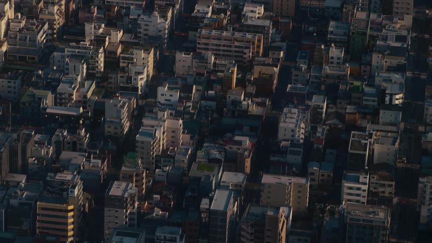 An aerial view captures the dense urban landscape of a city with shadows moving across the rooftops as the sun sets