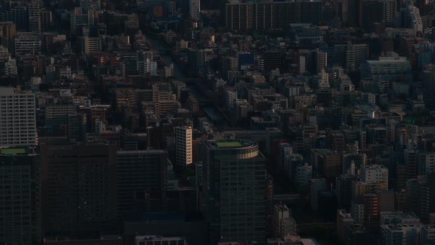 An aerial shot captures the densely packed buildings of a major city as the sun sets, casting long shadows across the urban landscape