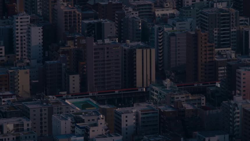 A commuter train travels on elevated tracks through a dense urban cityscape during a moody twilight