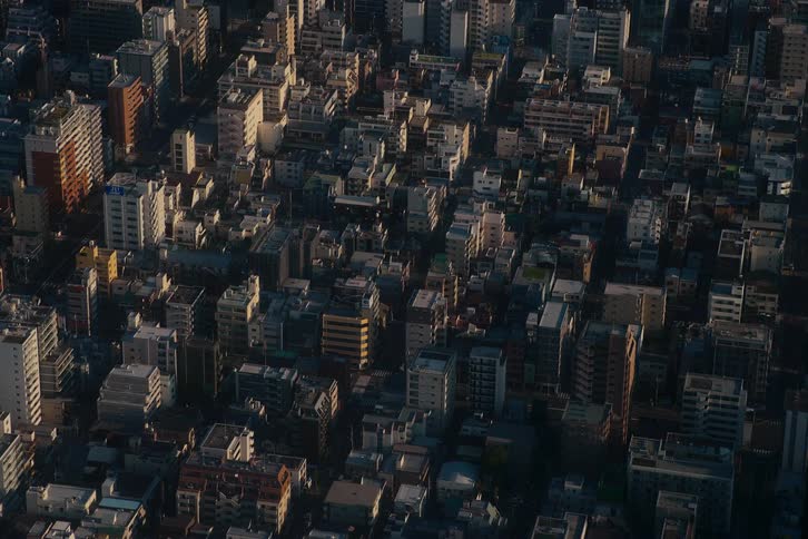 A static aerial view captures the sprawling, densely packed buildings of a city as golden morning light casts long shadows across the urban landscape