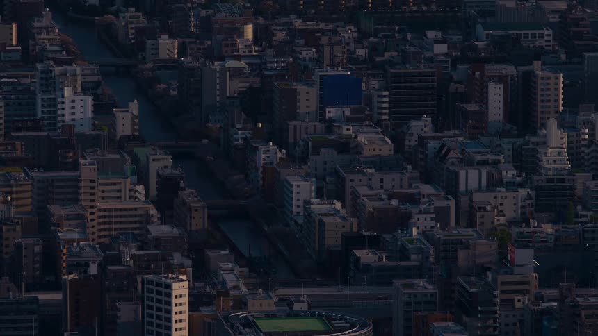A train travels through a dense urban cityscape in Japan as the setting sun casts long shadows across the buildings