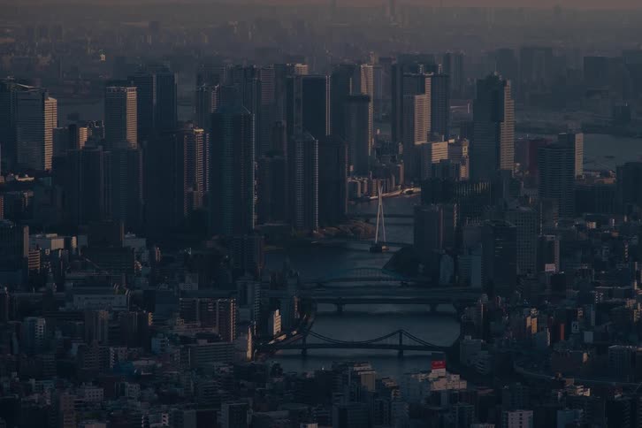 A moody, high-angle view of the Tokyo cityscape and the Sumida River as a lone boat travels through the hazy dusk