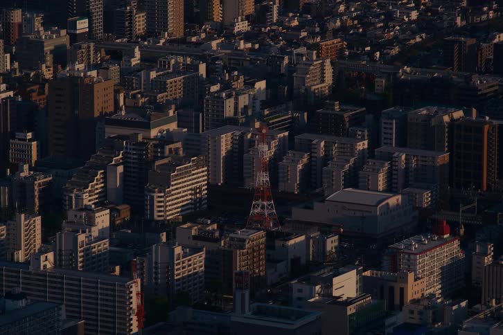 An aerial time-lapse captures the dense urban landscape of Kobe, Japan, as the setting sun casts long shadows across buildings surrounding the iconic Port Tower