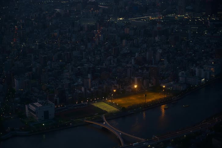 An aerial view shows a brightly lit park along a river contrasting with the sprawling, dark cityscape as night falls