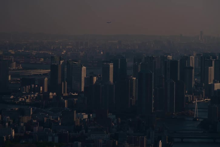 A static aerial shot of a dense, hazy cityscape at dusk as a lone airplane flies across the sky