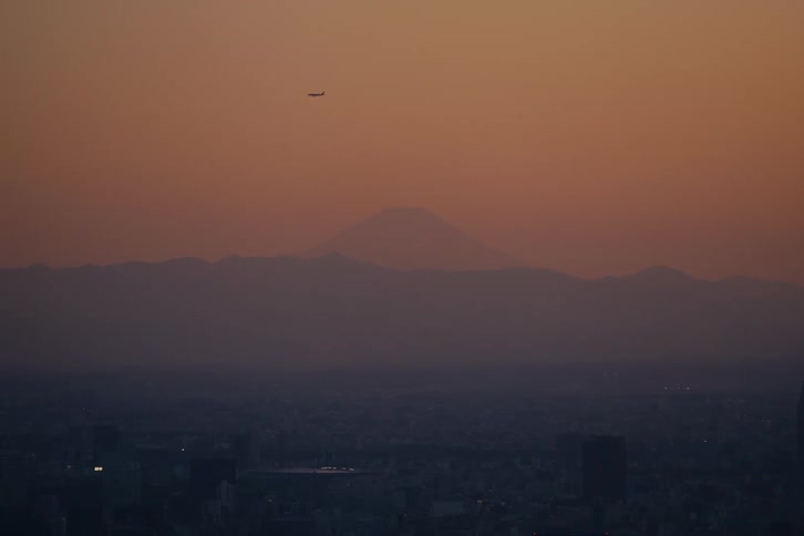 An airplane flies over a hazy Tokyo cityscape with the silhouette of Mount Fuji visible in the distance during a beautiful sunset