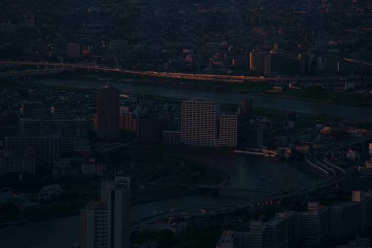 An aerial shot captures the last golden rays of sunset illuminating a distant highway in a vast, darkened city divided by a winding river