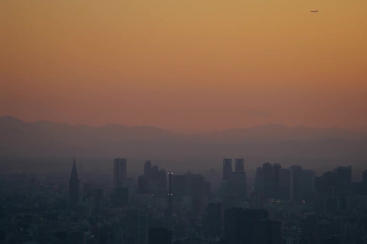 An airplane flies over a hazy city skyline silhouetted against a beautiful orange sunset sky with mountains in the distance