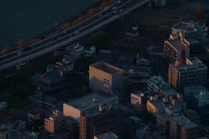 An aerial view of a Japanese neighborhood showing a traditional temple next to a busy highway along the water during a golden sunset