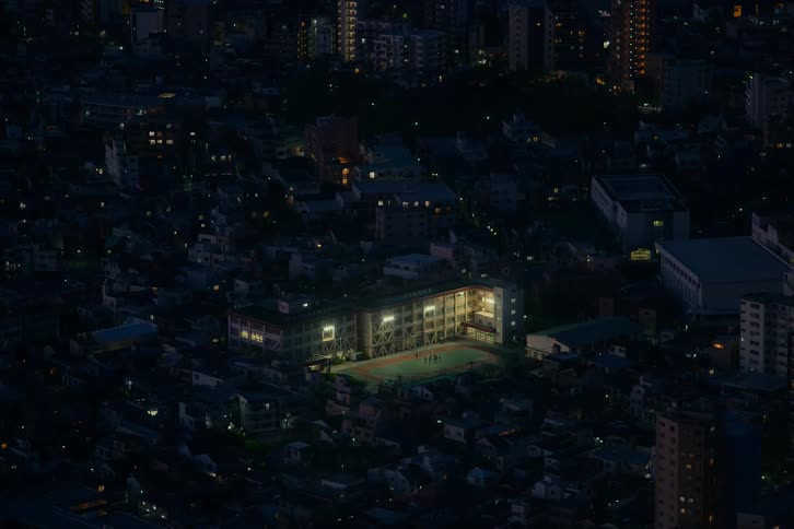 A high-angle view captures a brightly lit schoolyard, a lone beacon of activity where people play at night in a quiet, dark city