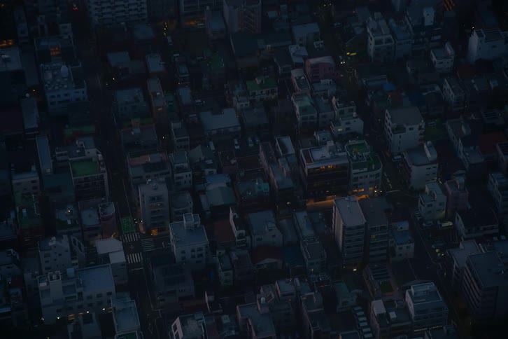 A high-angle view captures a dense urban neighborhood as lights begin to appear in the buildings at dusk