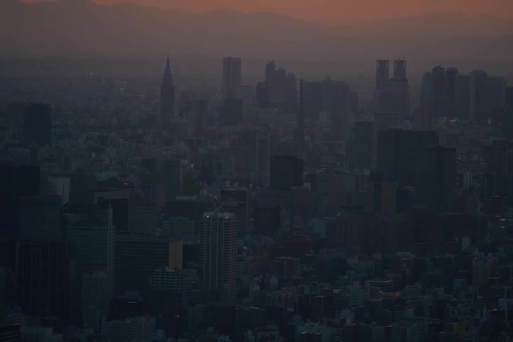 A moody, static view of a hazy urban skyline at dusk, with distant mountains silhouetted against the orange sky