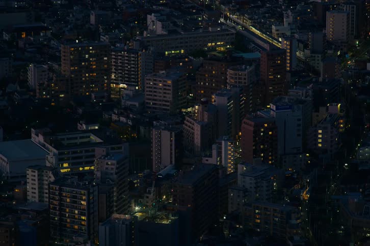 An elevated view captures the sprawling cityscape of a Japanese metropolis at night as a train quietly passes through the illuminated buildings