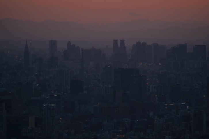 The hazy Tokyo skyline is silhouetted against a warm, colorful sunset with mountains in the distance