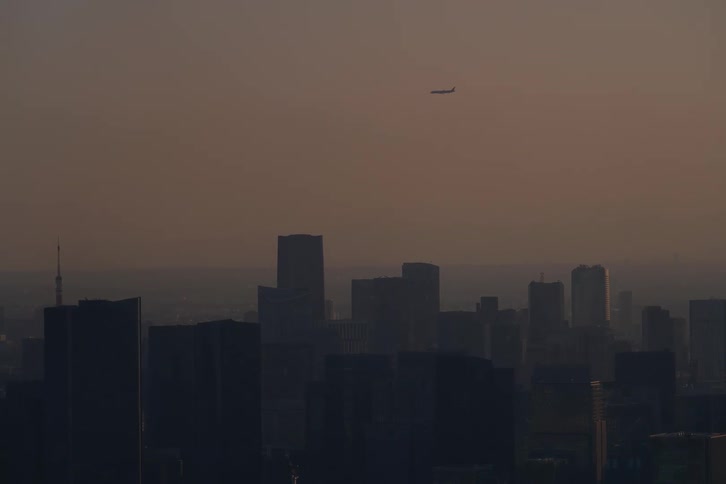 An airplane flies over a silhouetted cityscape during a hazy sunset