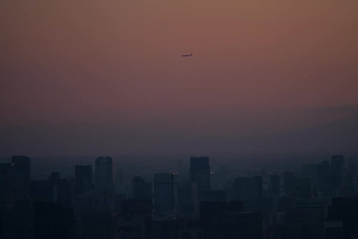 A lone airplane flies across the hazy sky over a metropolitan city during a serene sunset