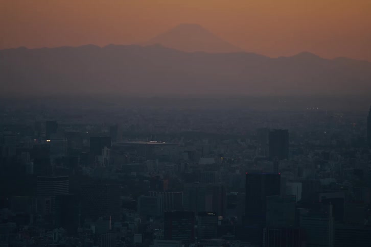 The iconic silhouette of Mount Fuji looms over the hazy Tokyo cityscape as the sun sets