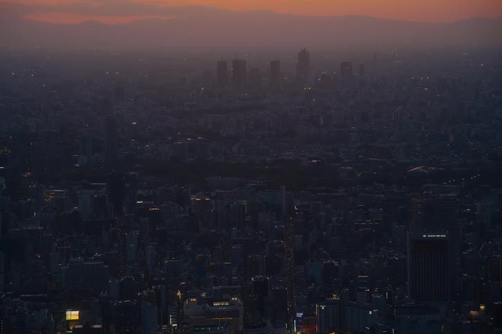 A vast and hazy cityscape of Tokyo fades into darkness as the sun sets, with skyscrapers silhouetted against the orange glow on the horizon