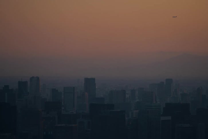 A lone airplane flies over a vast and hazy city skyline under the warm glow of a sunset