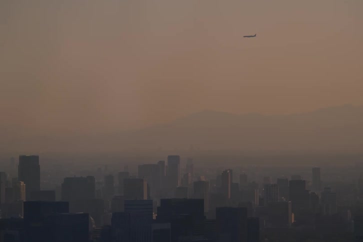 A passenger jet soars above a smog-filled city skyline during a hazy sunset