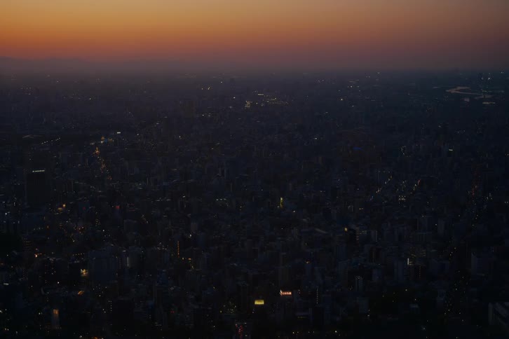 The last glow of an orange sunset fades over a sprawling cityscape as the urban area slowly comes to life with twinkling lights