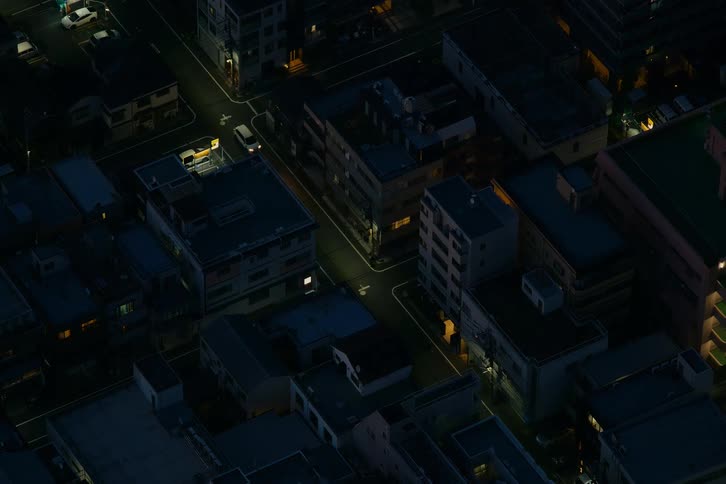 A high-angle view shows a single white car driving through a quiet, dimly lit urban neighborhood at night