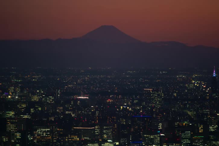 The iconic silhouette of Mount Fuji looms over the sprawling, illuminated Tokyo cityscape during a beautiful twilight