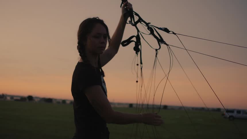 A female skydiver meticulously checks and sorts the lines of her parachute rig in a field during a golden hour sunset