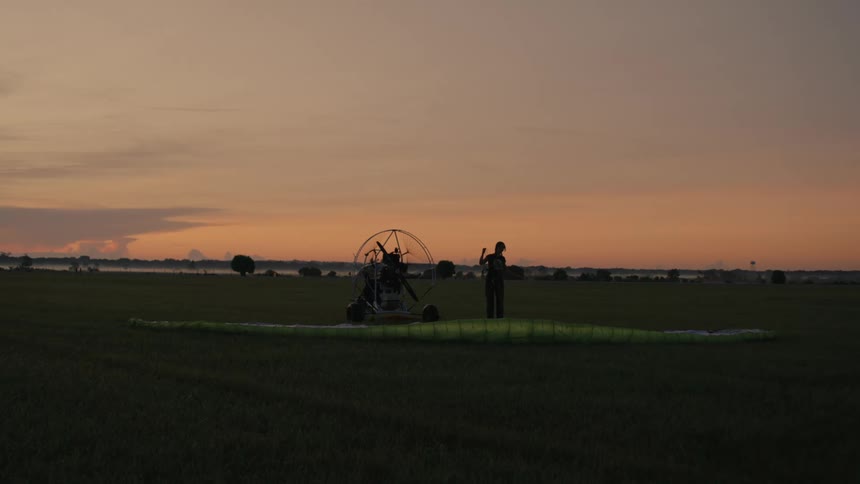 A silhouetted pilot prepares their powered paraglider for takeoff in a vast field at sunrise