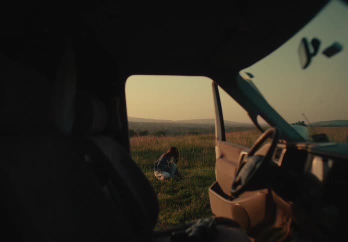 A woman is framed by an open car door as she peacefully picks wildflowers in a sun-drenched field at golden hour