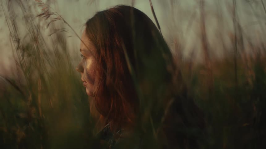 A thoughtful woman sits peacefully in a field of tall grass during a serene golden hour