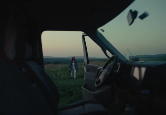 A serene view from inside a van of a woman standing in a beautiful, grassy landscape at twilight