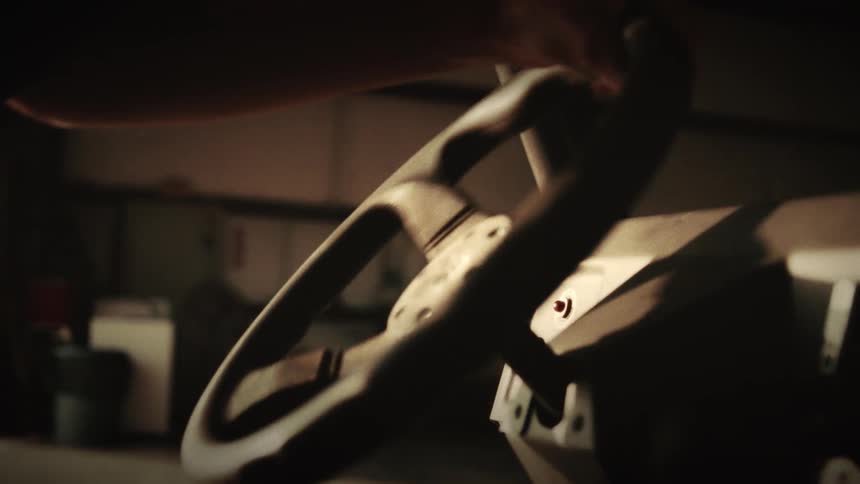A cinematic close-up shows a hand gripping and turning the steering wheel of a vehicle as it moves from a dark hangar into the daylight