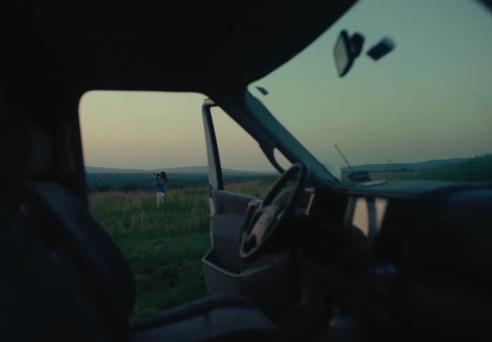 A cinematic view from inside a dark van captures a woman taking photos of the rolling hills at sunset