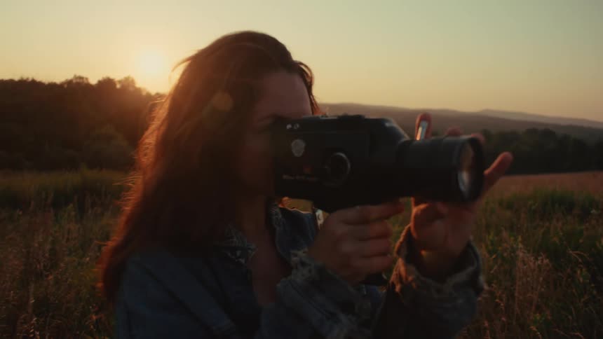 A woman with a vintage film camera captures the beautiful golden light of sunset in a grassy field