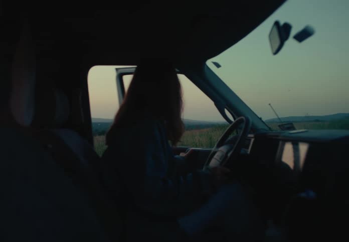 A silhouetted woman sits in the driver's seat of a van, parked in a rural field during a serene twilight