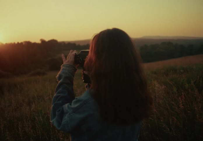 A young woman stands in a grassy field at golden hour, filming the sunset with a vintage movie camera