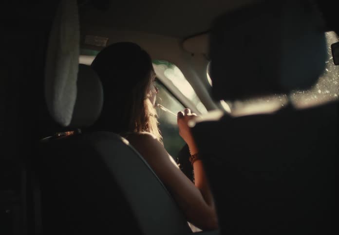 A woman sits in the driver's seat of her car applying makeup as the warm glow of the sun shines through the window
