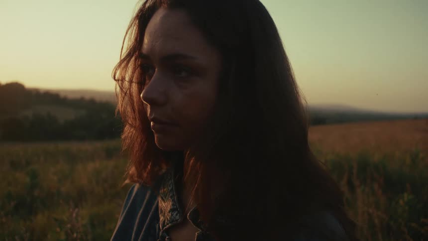 A melancholic young woman looks away pensively while standing in a sunlit field at dusk