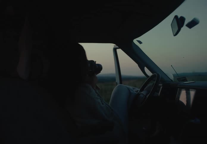 A person sits peacefully in a van filming the quiet twilight landscape with a vintage video camera