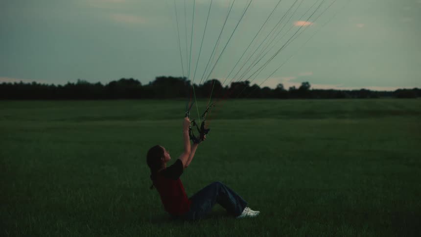 A young woman paraglider sits in a grassy field at twilight, holding the risers as her canopy inflates in the wind