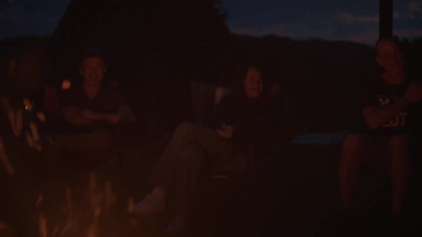 A group of friends laugh and chat together in slow motion as they sit around a crackling campfire at dusk