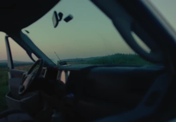 A dreamy, slow pan from inside a parked van reveals a person taking photos of the beautiful countryside landscape at sunset