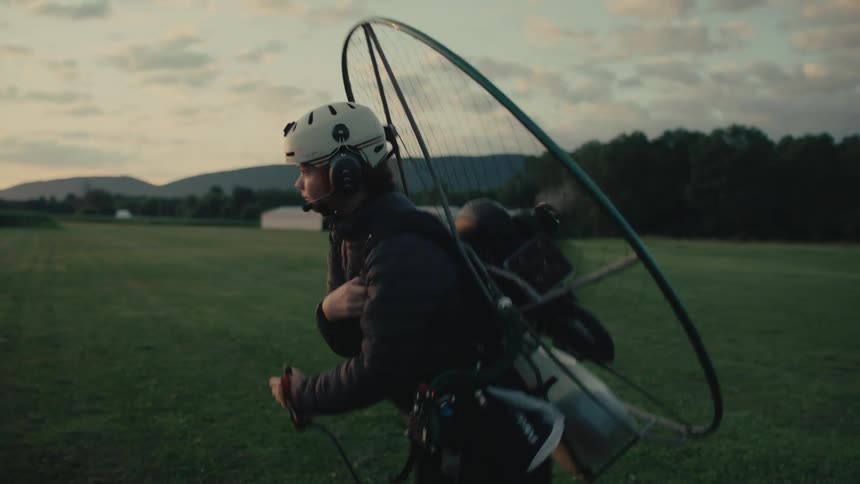 A pilot wearing a helmet starts the engine of his powered paraglider in a grassy field at dusk, the propeller spinning in slow motion