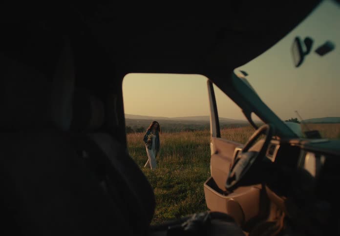 View from inside a car of a young woman picking wildflowers in a grassy field during a golden hour sunset