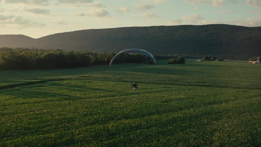 An aerial view shows a powered paraglider running across a vibrant green field to take off at sunset