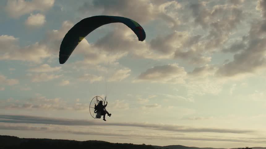 A silhouetted powered paraglider soars peacefully through a cloudy sky during a warm, golden sunset