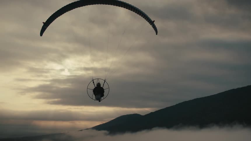 A silhouetted powered paraglider floats peacefully above a sea of clouds as the sun sets behind the mountains