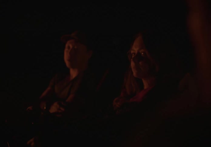 A couple sits in chairs by a flickering campfire at night, their faces dimly lit by the flames