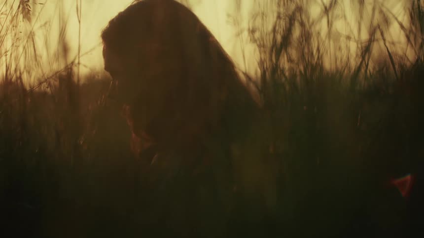 Close up of a thoughtful woman's silhouette as she sits amongst tall grass at sunset